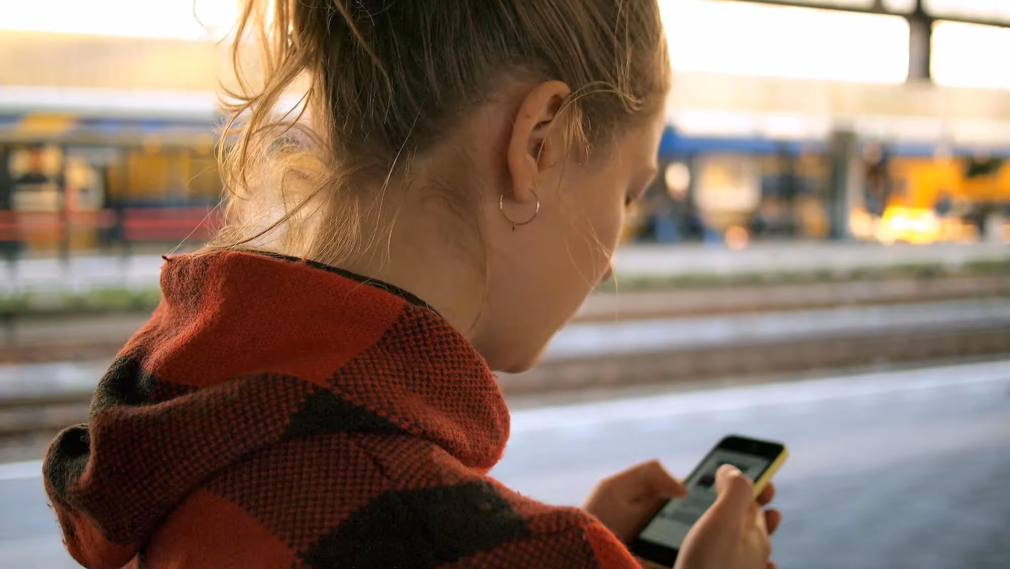 Woman in a red and black checkered hoodie looking at her smartphone at a train station platform.