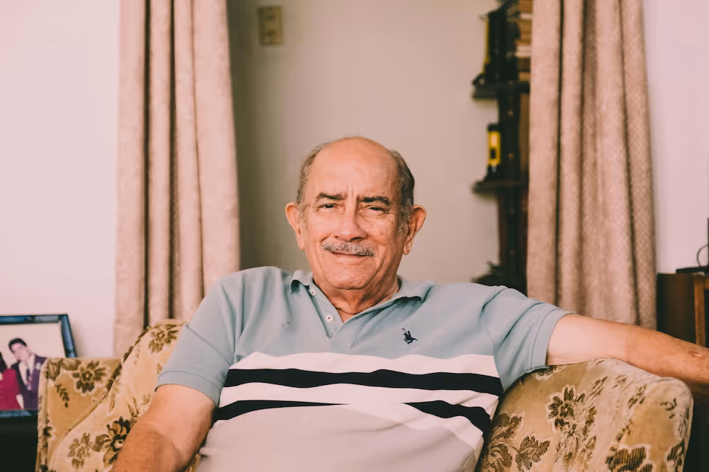 Smiling elderly man sitting on a beige floral-patterned couch in a living room.