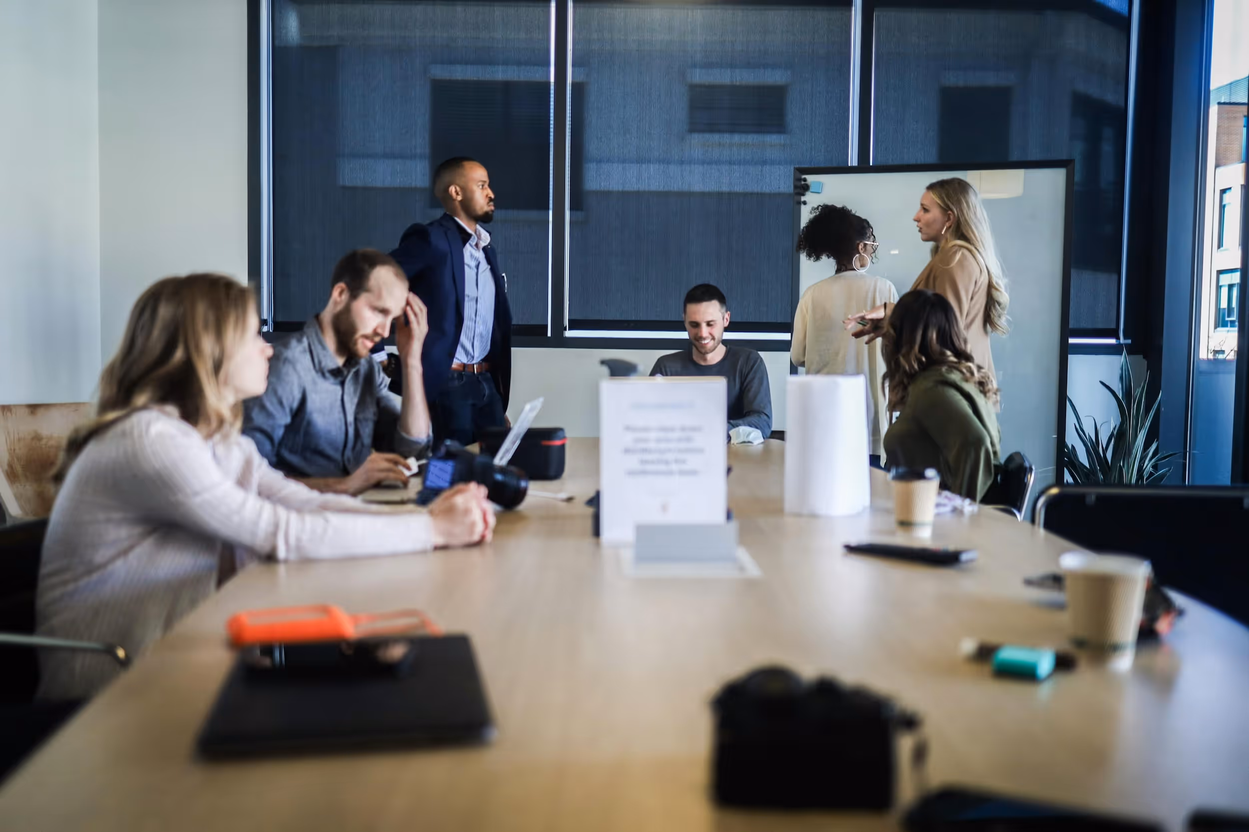 Group of six people having a meeting in a modern office with some standing near a whiteboard and others seated around a conference table.