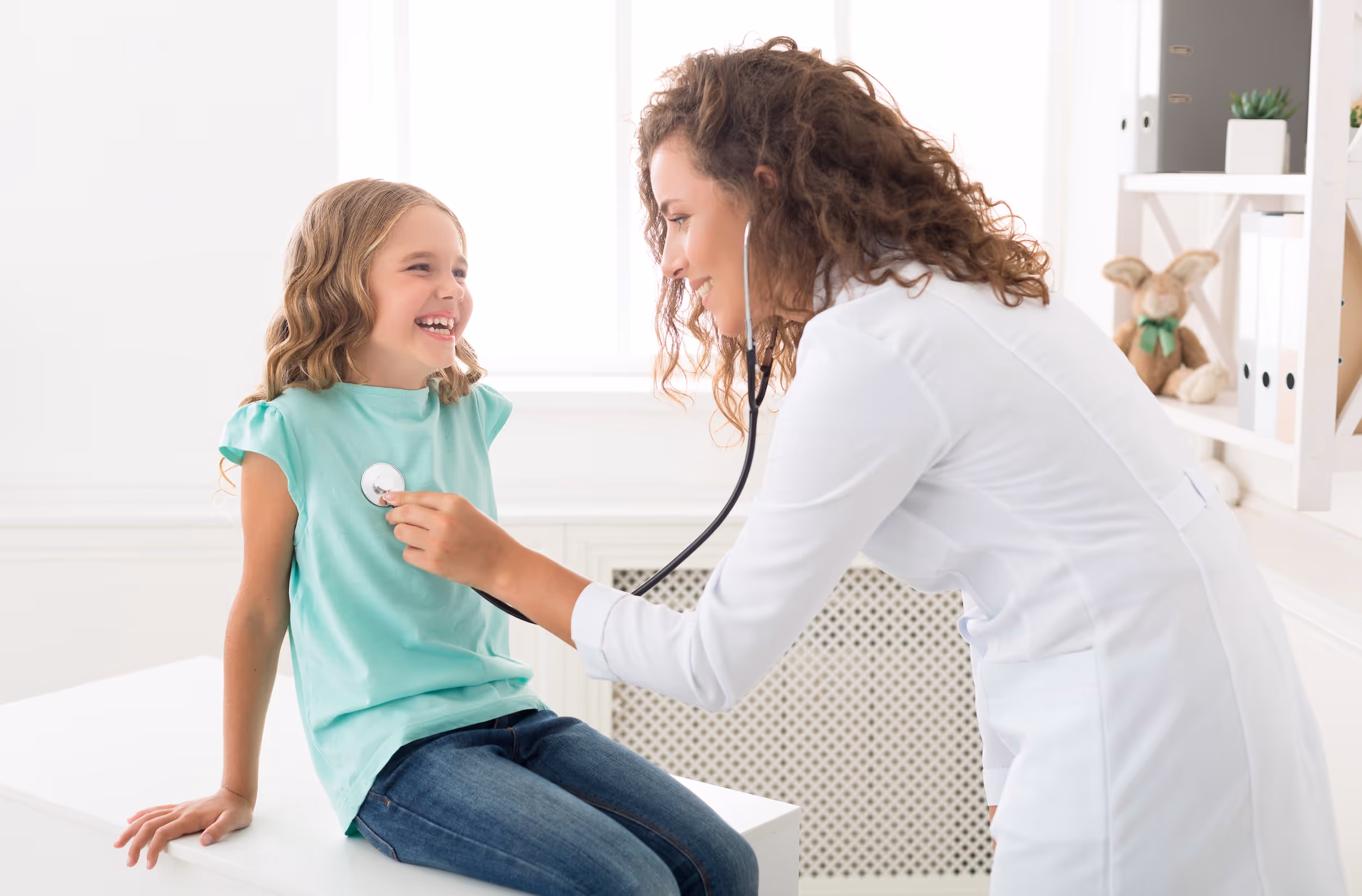 Female doctor using a stethoscope to listen to a smiling young girl's chest in a bright medical office.