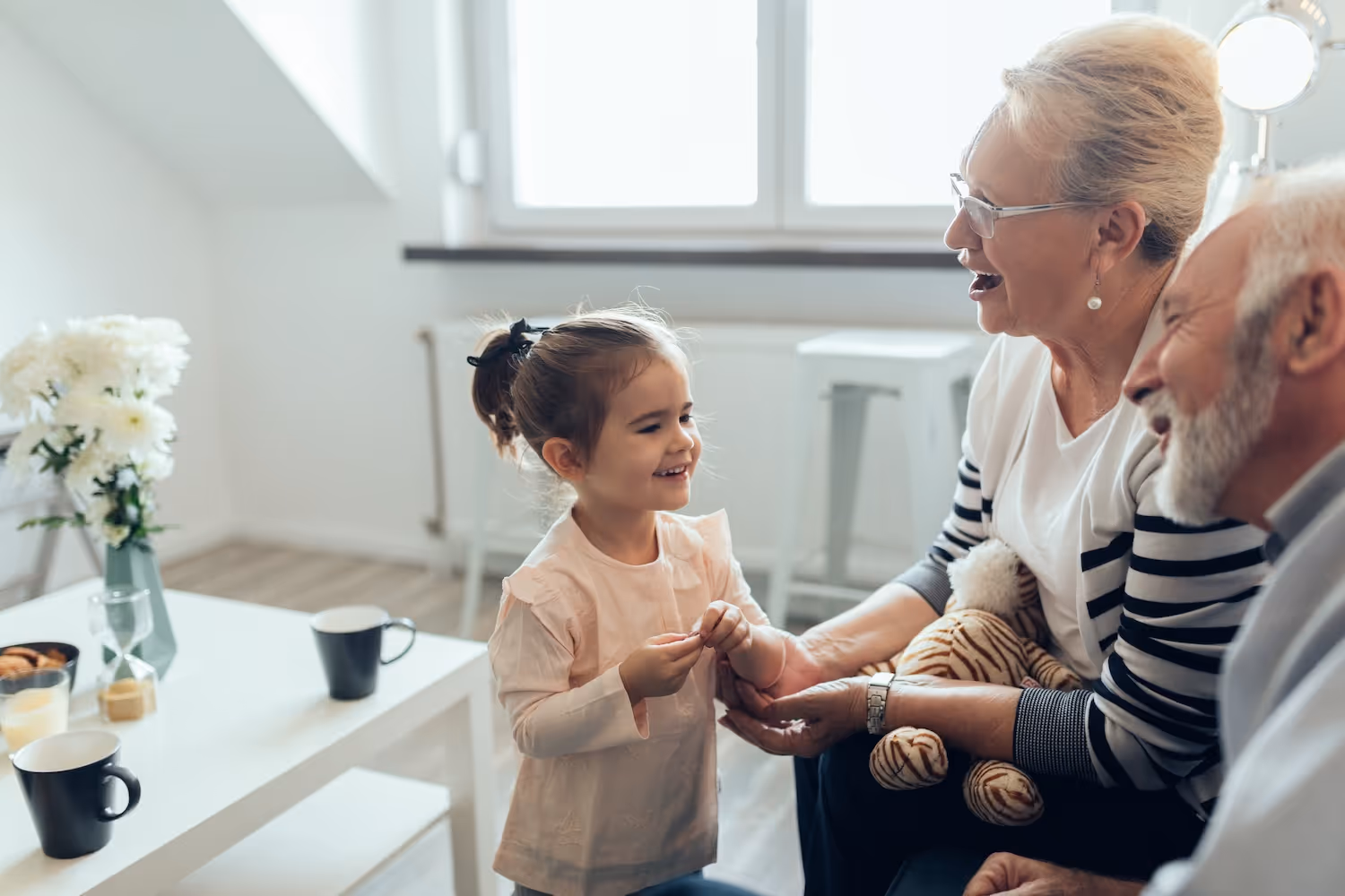 Smiling young girl holding hands with her grandmother while sitting next to her grandfather in a bright living room.