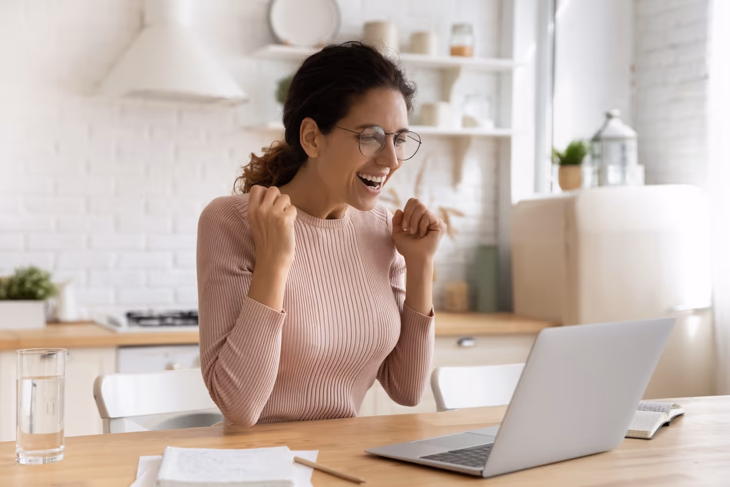 Smiling woman wearing glasses celebrating while looking at a laptop in a bright kitchen.