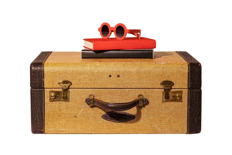 Vintage Reisekoffer mit braunem Griff und Verschlüssen, darauf zwei Bücher und eine rote Sonnenbrille.