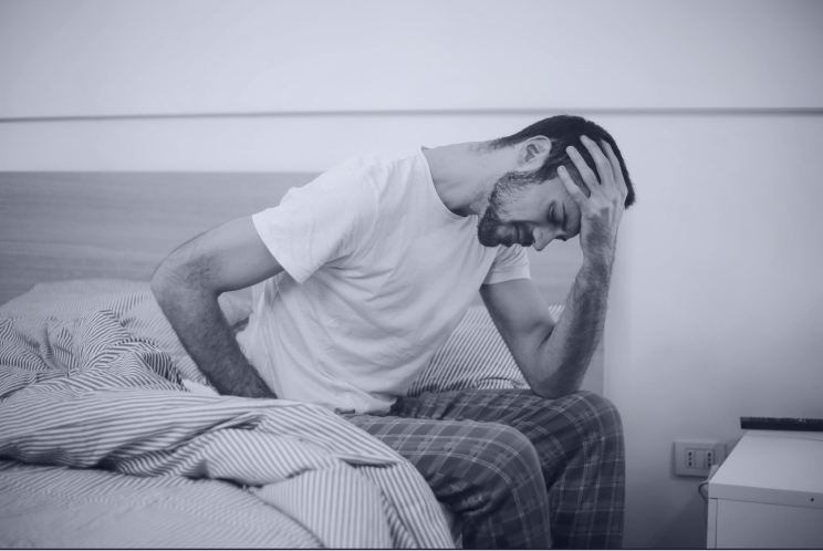 A man sitting in his bed with pijamas, he is holding his face with a sad face as if the has a headache