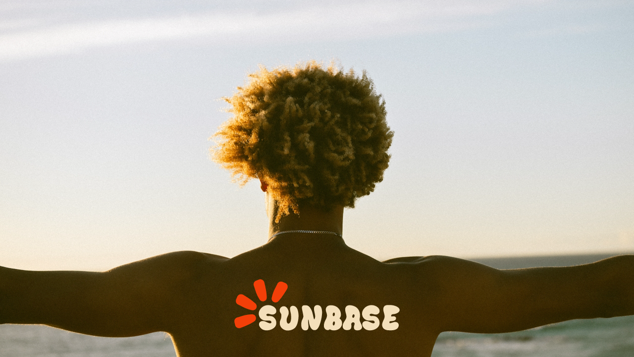 Back view of a shirtless person with outstretched arms and curly hair at the beach with Sunbase logo.