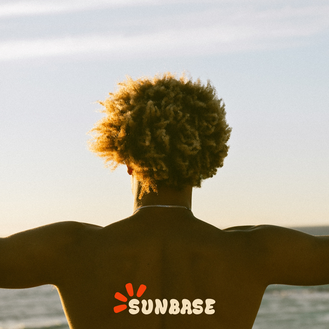 Back view of a shirtless person with curly hair standing with arms outstretched by the sea at sunset.
