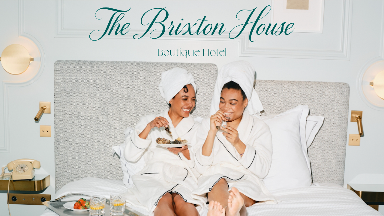 Two women in white robes and towels on their heads relax on a hotel bed with snacks and drinks.