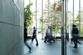 Employees move quickly in a bright building with tall windows.