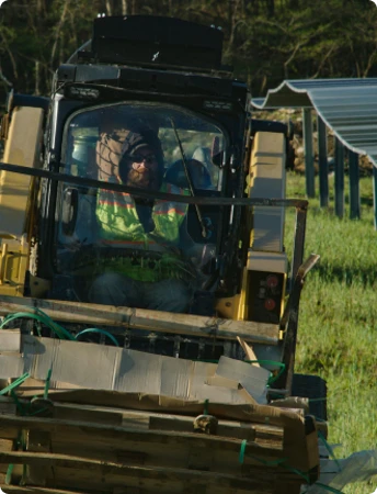 Skid Steer Loader Transporting Materials on Farm Site Operator driving a skid steer loader carrying wood pallets and pipe materials across a farm field, with a hoop house structure in the background
