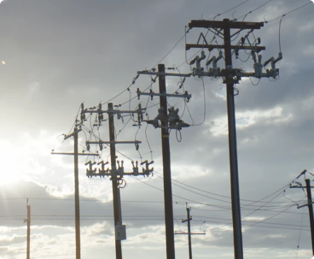 Utility power poles with electrical transformers and crossarms against a cloudy sky, backlit by sunlight