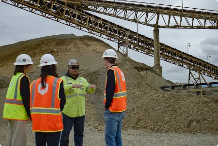 Site Team Discussion at Aggregate Facility Team of four construction workers in hard hats and safety vests discussing project details at an aggregate mining site with conveyor structure
