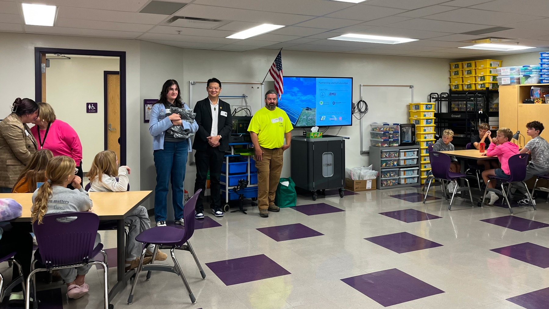 Project partners stand at the front of a Bryan Elementary STEM classroom beside a screen showing a solar array and partner logos, as students look on.