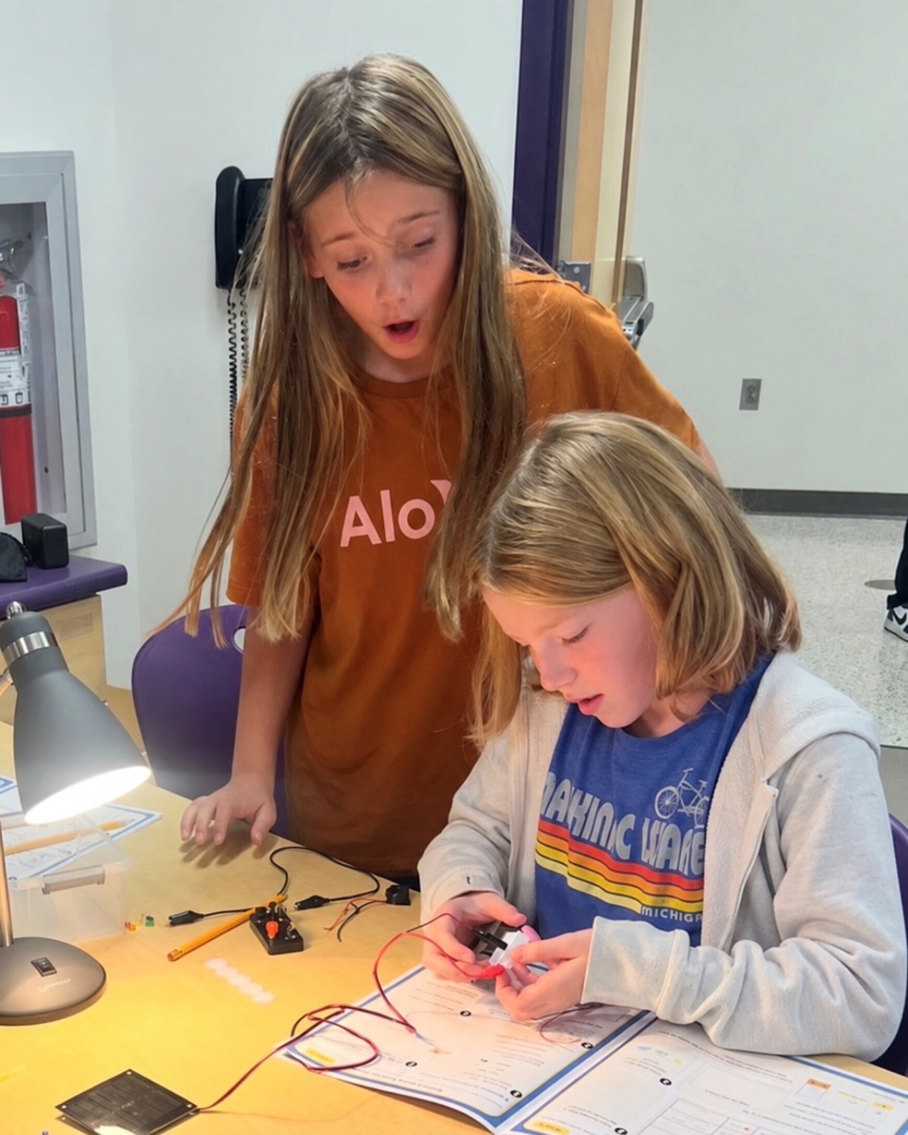 A student connects wires to a small motor while a classmate reacts with surprise, with a solar panel, desk lamp, and activity workbook on the table.