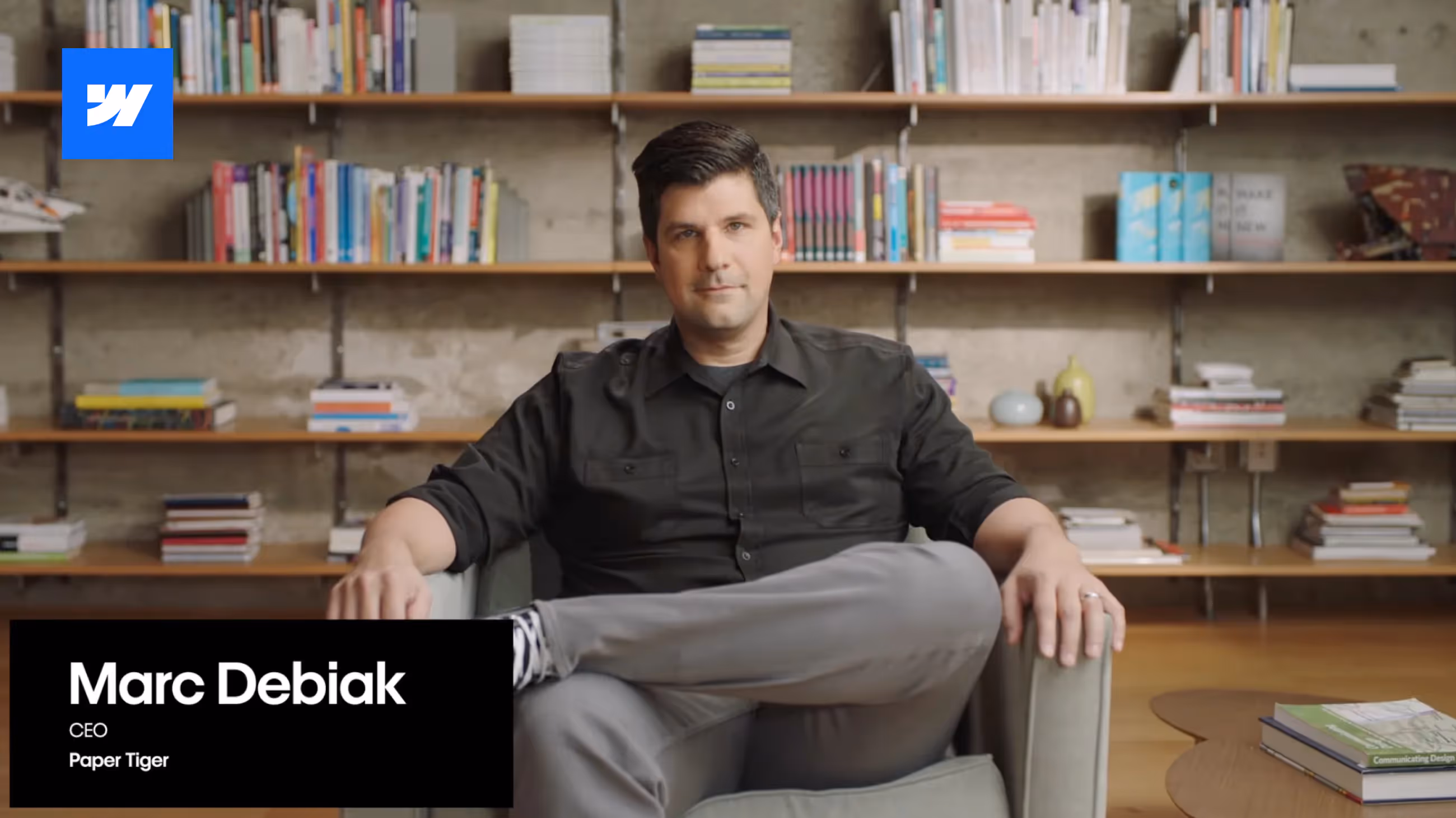 Screenshot of Webflow video:Paper Tiger's CEO Marc Debiak sitting on a chair with legs crossed, in front of a library wall