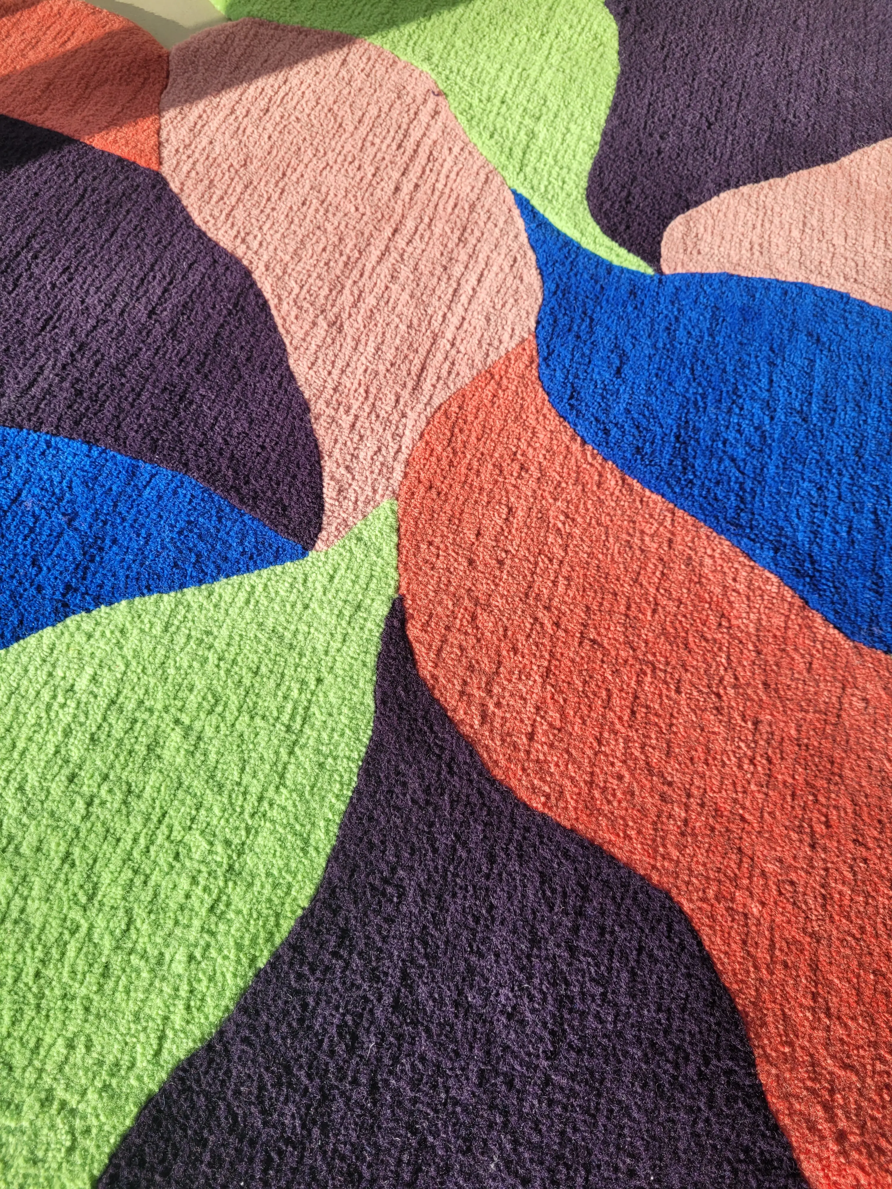 Hand-tufted colorful rug titled “Vague” with bold organic shapes in red, pink, green, blue, and purple, placed in a sunlit room.