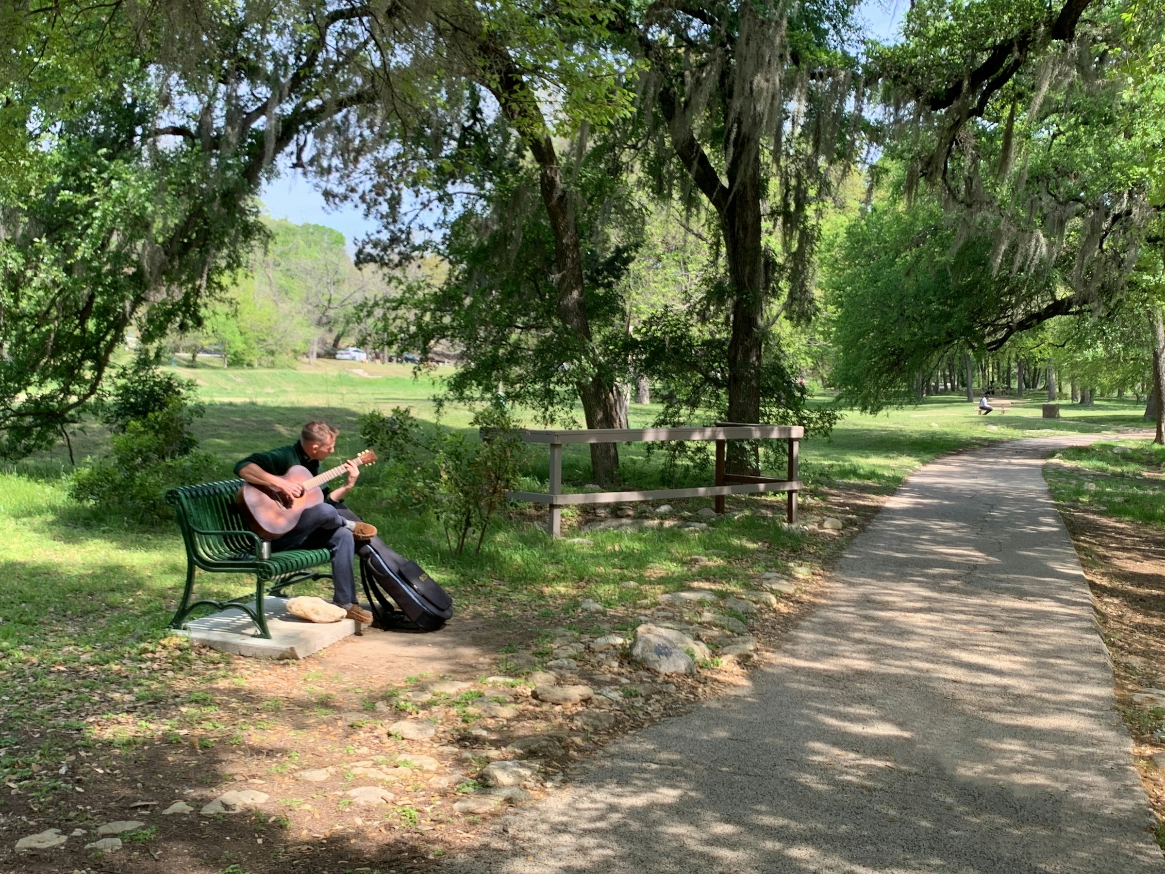 A man playing guitar on a park bench under the trees in Old West Austin