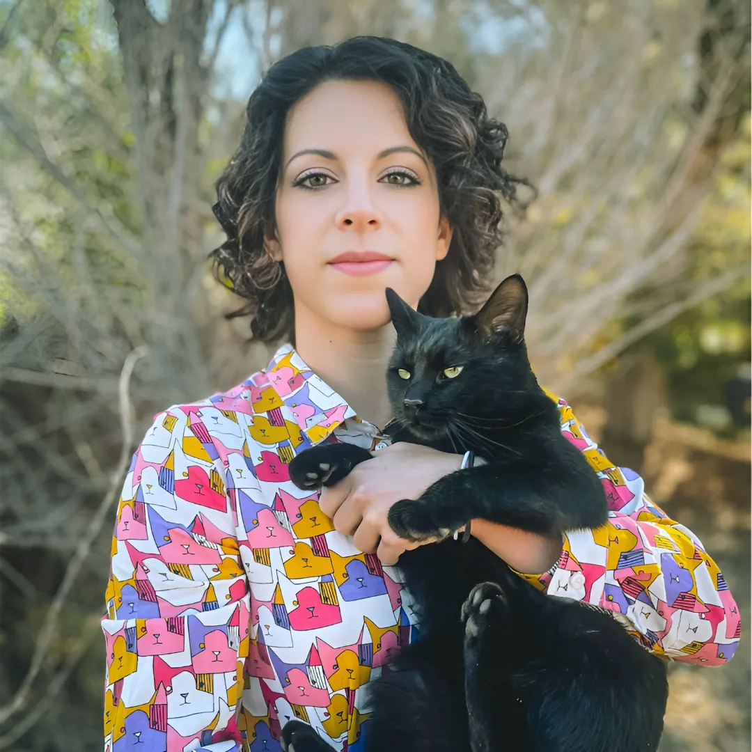 Woman with short curly hair wearing a colorful shirt with cat faces pattern holding a large black cat outdoors.