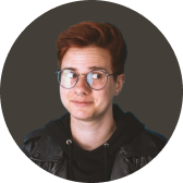 Person with short red hair, glasses, and a black hoodie under a leather jacket against a dark background.