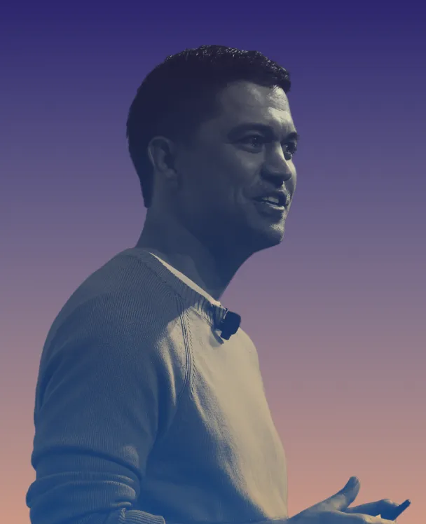 Man in a sweater speaking with a microphone clipped on his collar against a gradient purple to peach background.