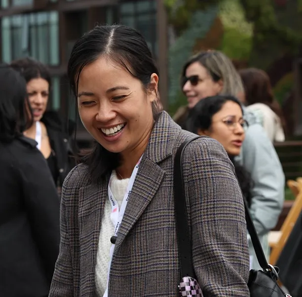 Smiling woman in a brown plaid blazer with black shoulder bag in an outdoor social setting.