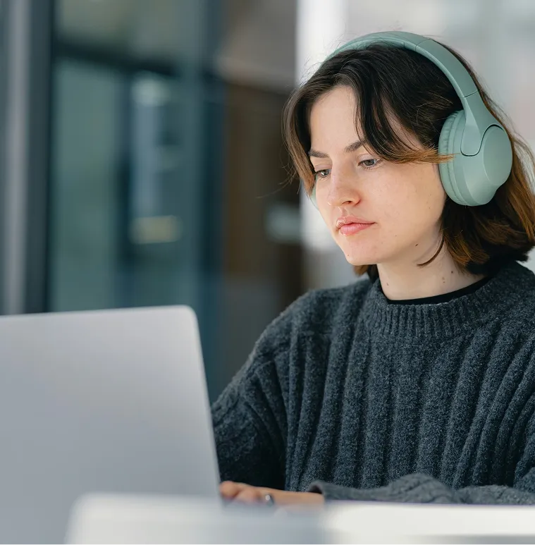 Young woman wearing green headphones and a dark gray sweater working on a laptop indoors.