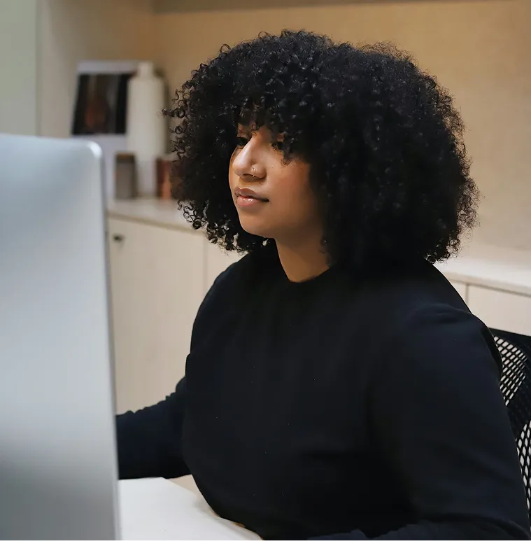 Woman with curly hair and nose ring sits at a desk working on a computer.