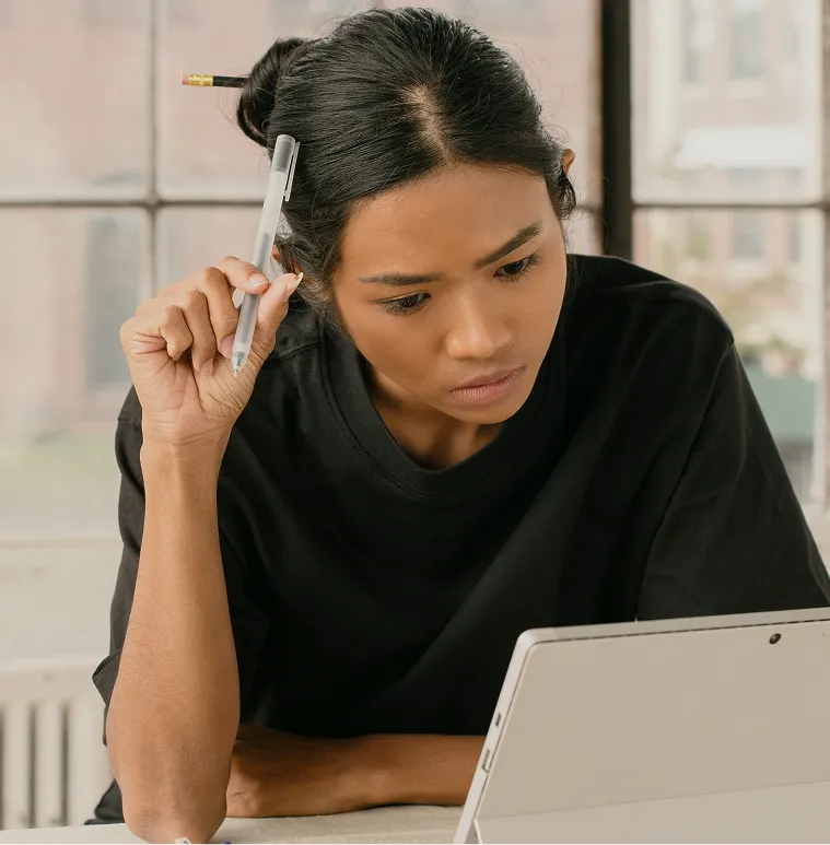 Woman with a pencil in her hair and holding a pen intently looking at a tablet.
