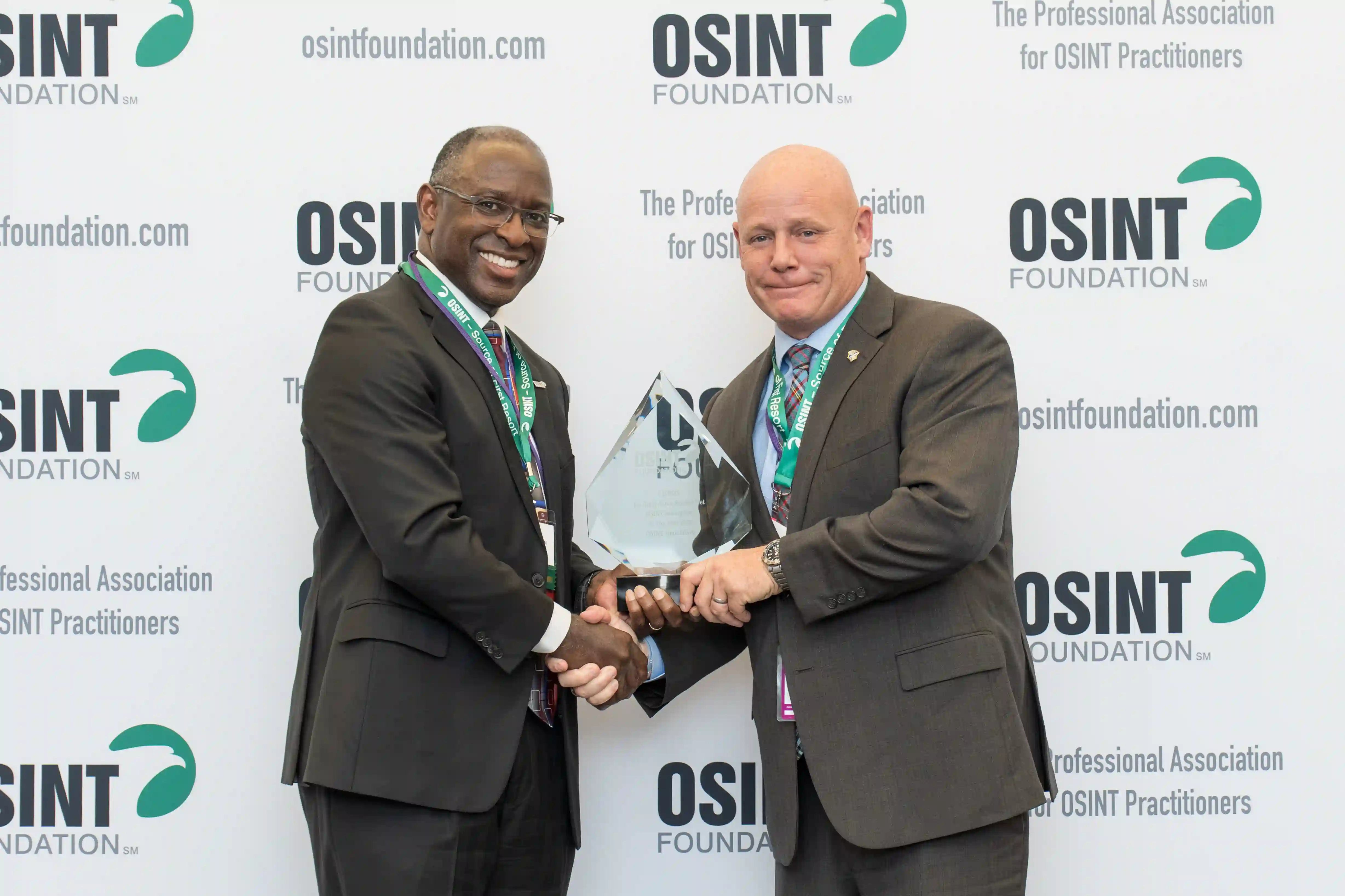man giving award to another man and shaking hands in front of step and repeat banner