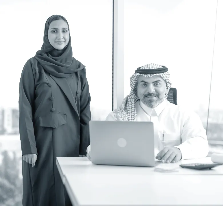 Two Middle Eastern business professionals in traditional attire in a modern office, one seated at a laptop and the other standing beside him.