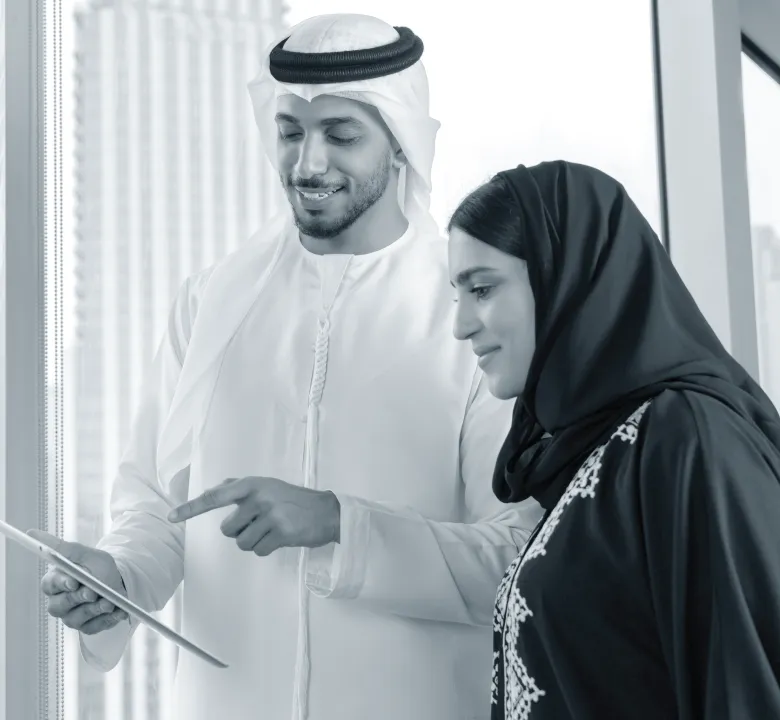Man in traditional Middle Eastern attire showing a tablet to a woman wearing a hijab, both standing near a window.