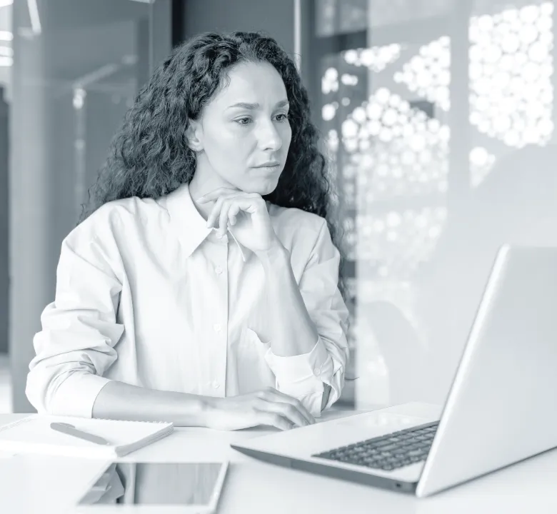 Woman with curly hair sitting at a desk, looking thoughtfully at a laptop screen with a notebook and tablet nearby.