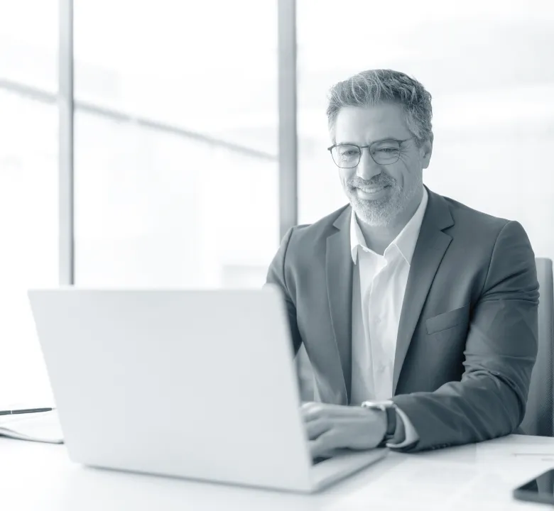 Middle-aged man with glasses in a suit smiling while working on a laptop in a bright office.