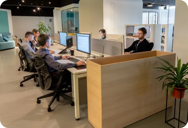 Office workspace with multiple people working at desks with computer monitors and a large wooden planter with a green plant.