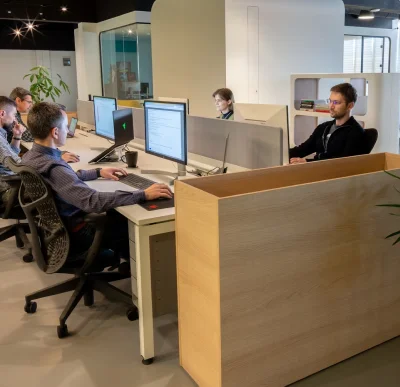 Office scene with four people working at desks with computer monitors, surrounded by partition walls and plants.