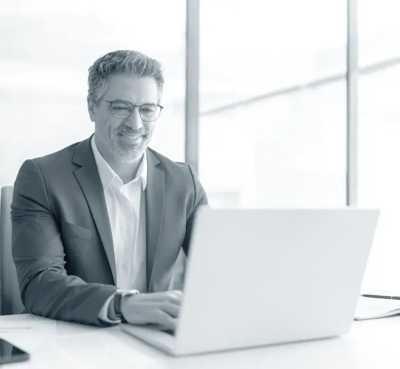 Smiling middle-aged man in glasses and suit working on a laptop at an office desk.