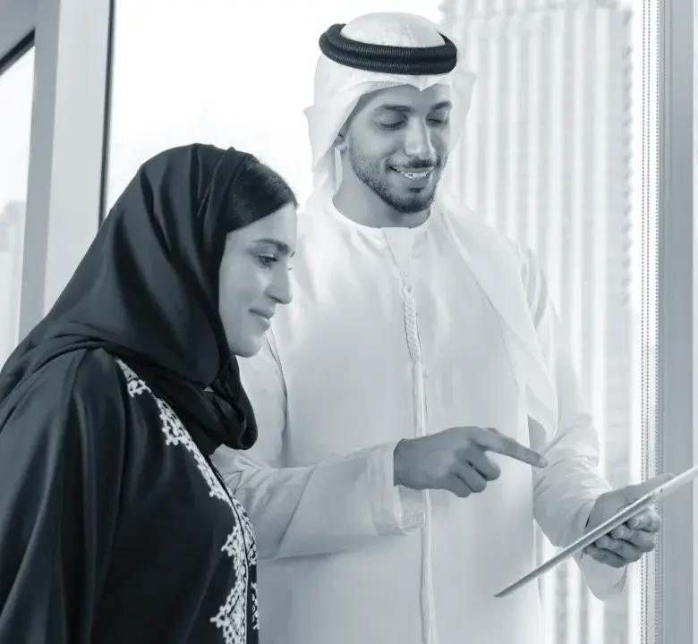 A man in traditional Middle Eastern attire showing a tablet to a woman wearing a hijab, both smiling and standing by a window in an office setting.