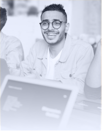 Young man with glasses smiling and sitting at a table with a laptop in front of him.