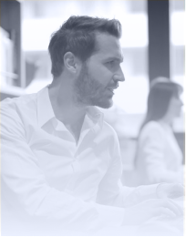 Side profile of a man with short hair and beard wearing a white shirt, speaking in an office setting with a blurred person in the background.