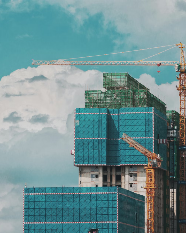 High-rise building under construction with blue scaffolding and yellow cranes against a cloudy blue sky.