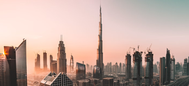Dubai city skyline at sunset with the Burj Khalifa towering in the center surrounded by skyscrapers and construction cranes.