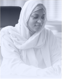 Woman wearing a white hijab and blouse, seated and looking down with a thoughtful expression.