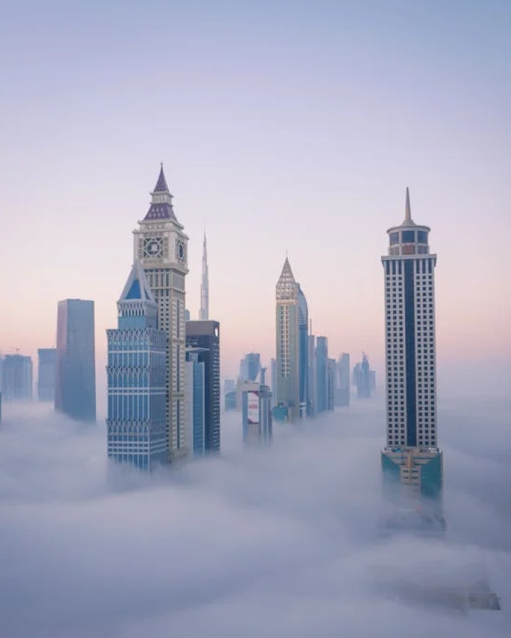 Skyscrapers emerging through dense fog in a cityscape at dawn with soft pastel sky.