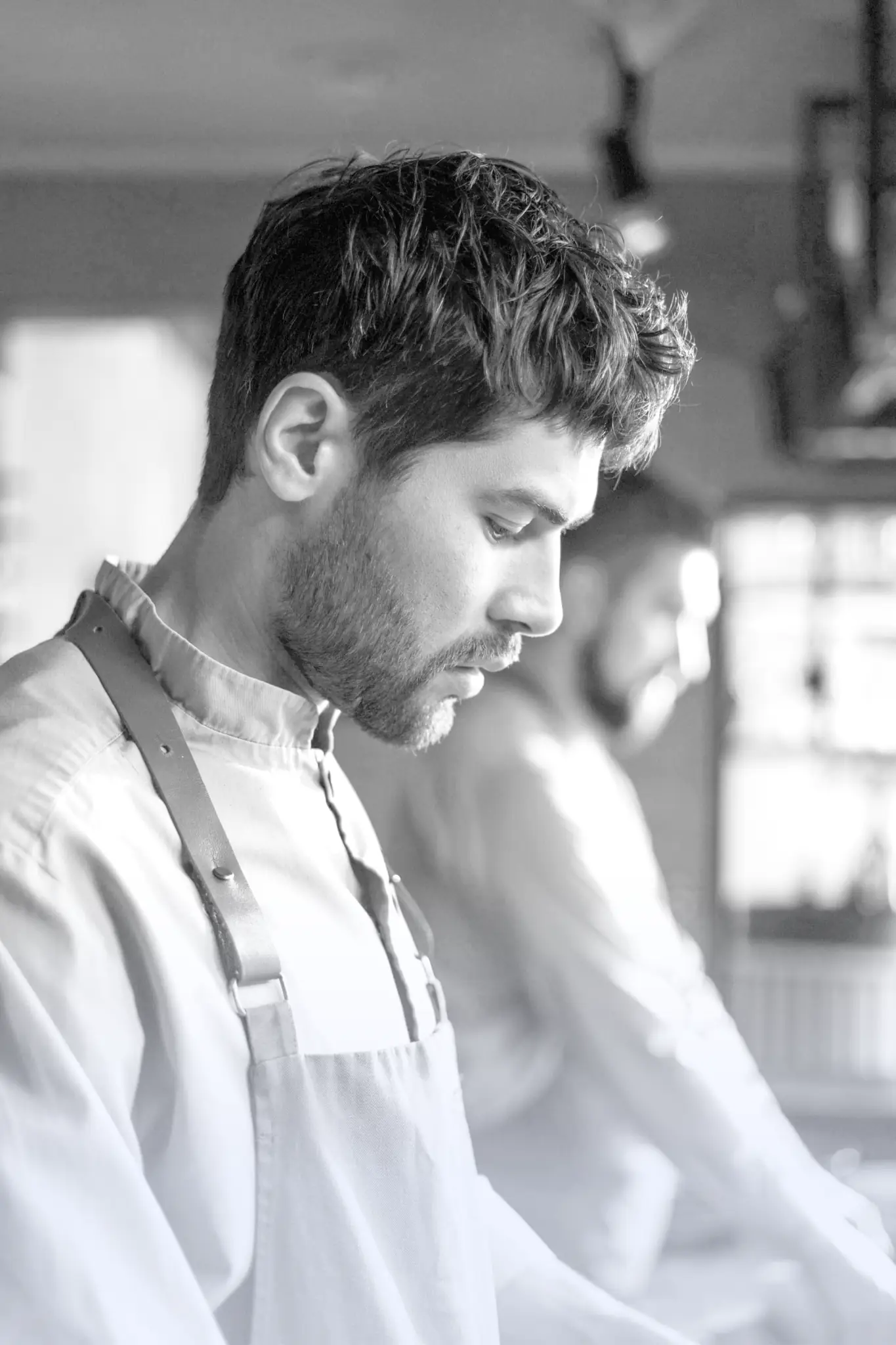 Close-up of a focused male chef wearing a grey apron with leather straps, working in a dimly lit kitchen.