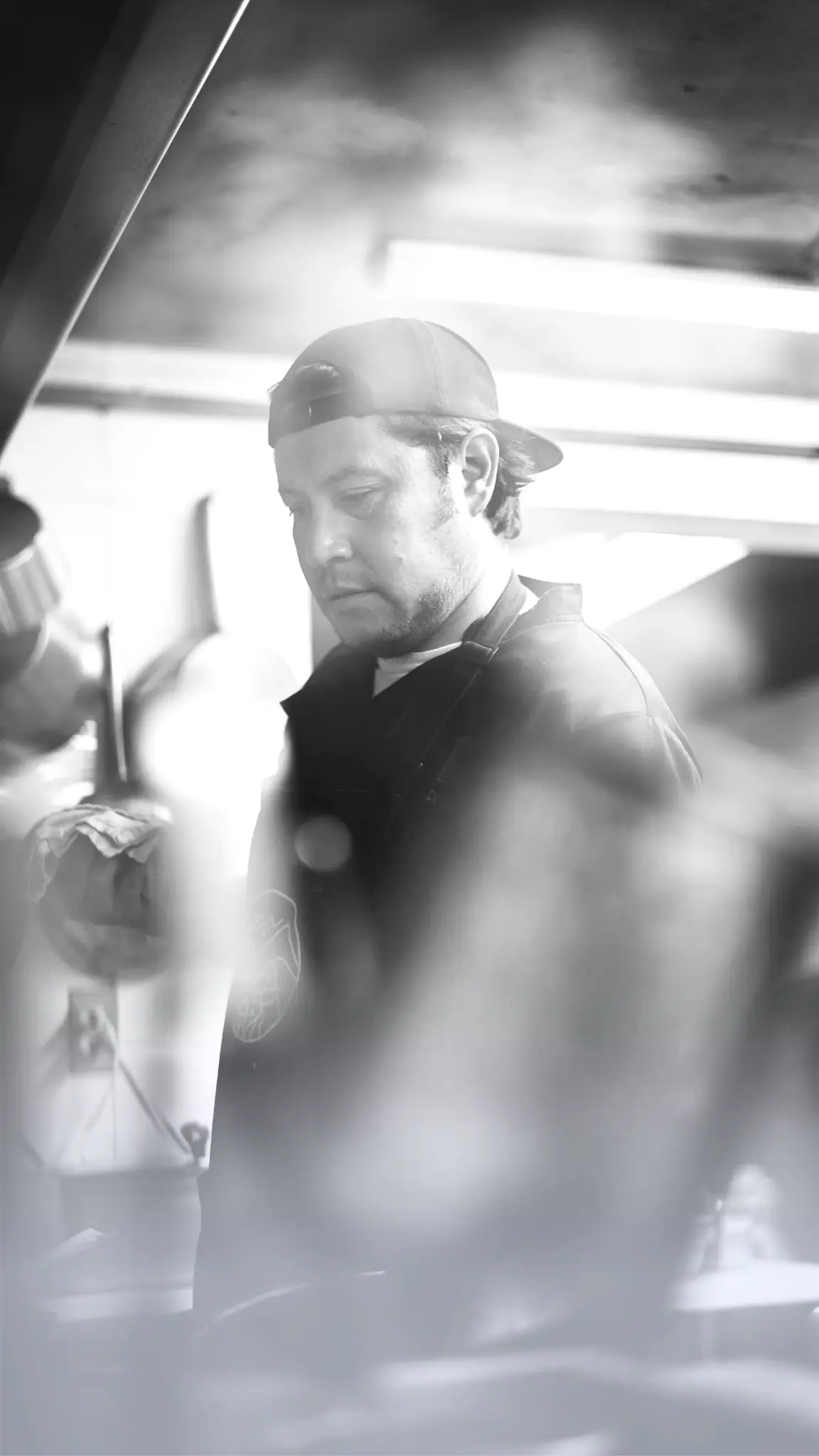 Man wearing a backward black cap and dark apron working in a kitchen with a focused expression.