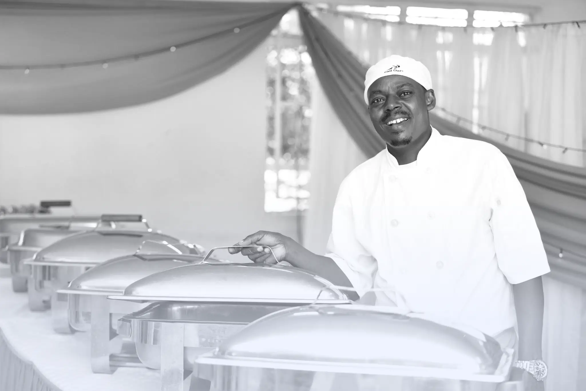 Chef in white uniform lifting lid of stainless steel chafing dish at a buffet table with gold and white drapery background.