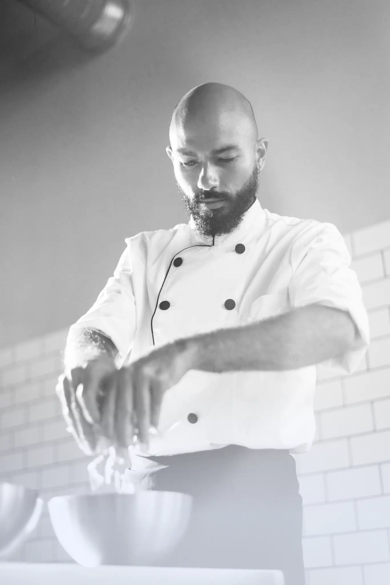 Chef in white uniform preparing food with fresh greens over a metal bowl in a kitchen.