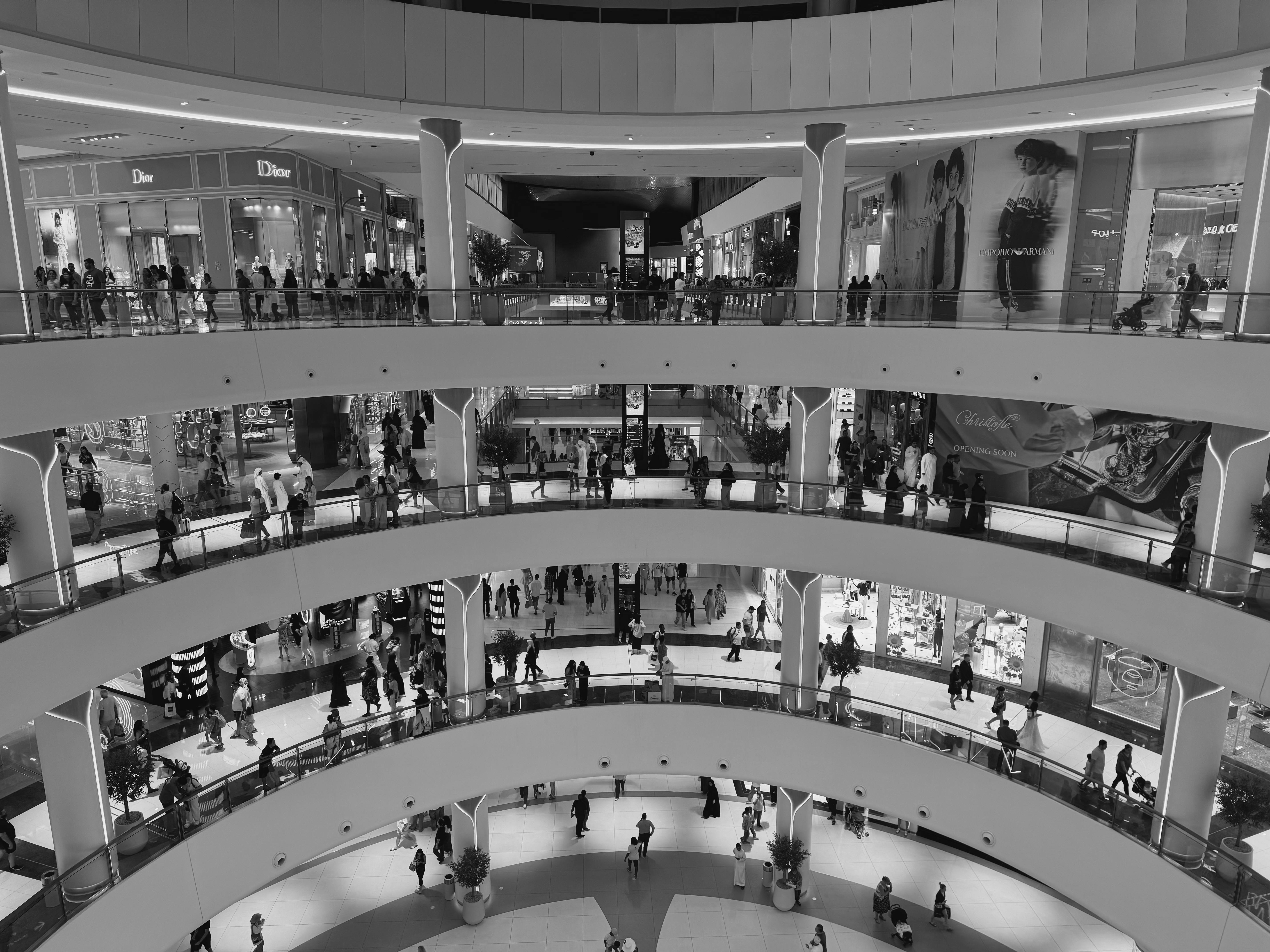 Overhead view of a multi-level shopping mall with people walking on various floors and store signs including Dior visible.