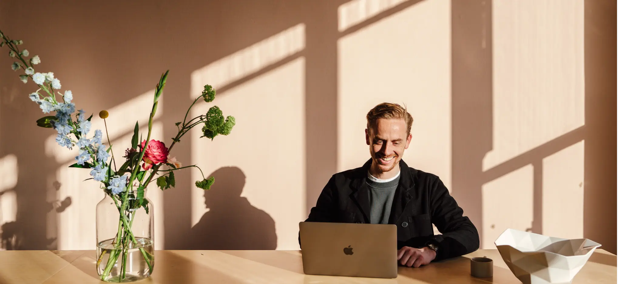 Man working on a laptop at a wooden table in a bright, sunlit home interior.