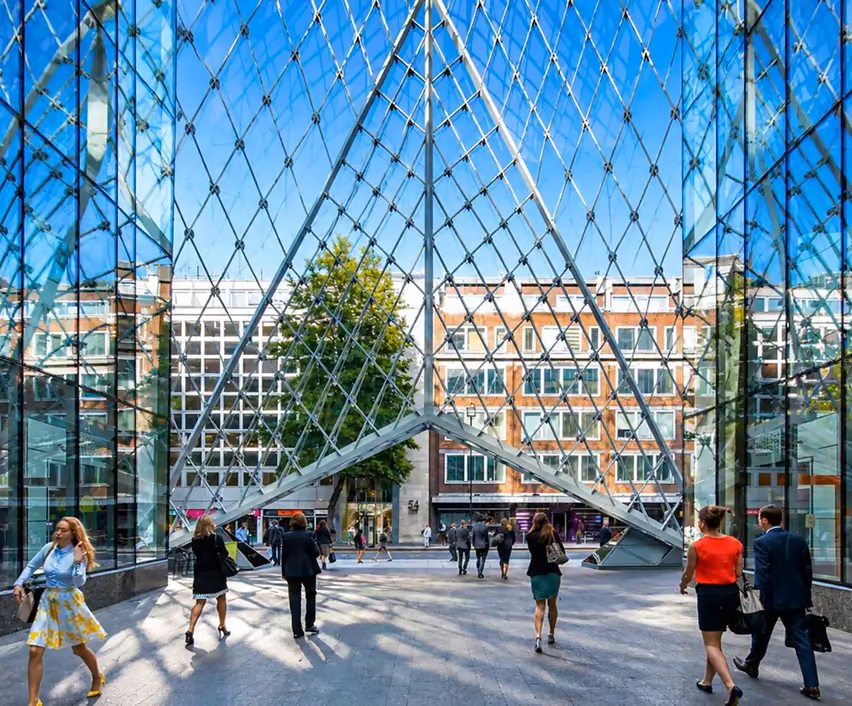 Office workers crossing a sunlit plaza beneath a diamond-lattice glass atrium