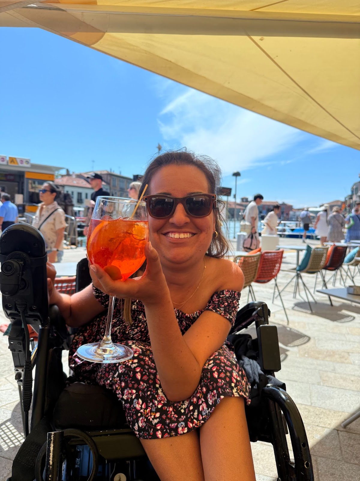 Smiling woman in a wheelchair wearing a dress and drinking an Aperol Spritz